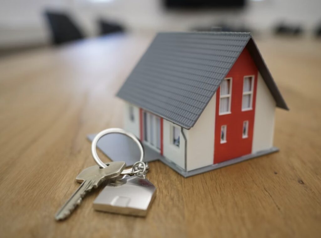 Close-up of a miniature house with a key and house-shaped keychain on a wooden table, symbolizing real estate or homeownership.